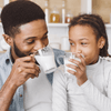 A father and daughter sitting in the kitchen drinking glasses of goat milk.