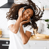 A smiling women in her 30s with curly brown hair and a white teeshirt listing to music with noise cancelling headphones and moving her head from side to side so that her hair is flying through the air.