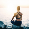 A women sitting on a rock on the side of a lake looking away from the camera at the sunset. She is wearing black active wear and meditating in the legs crossed position with her hands resting on her knees.